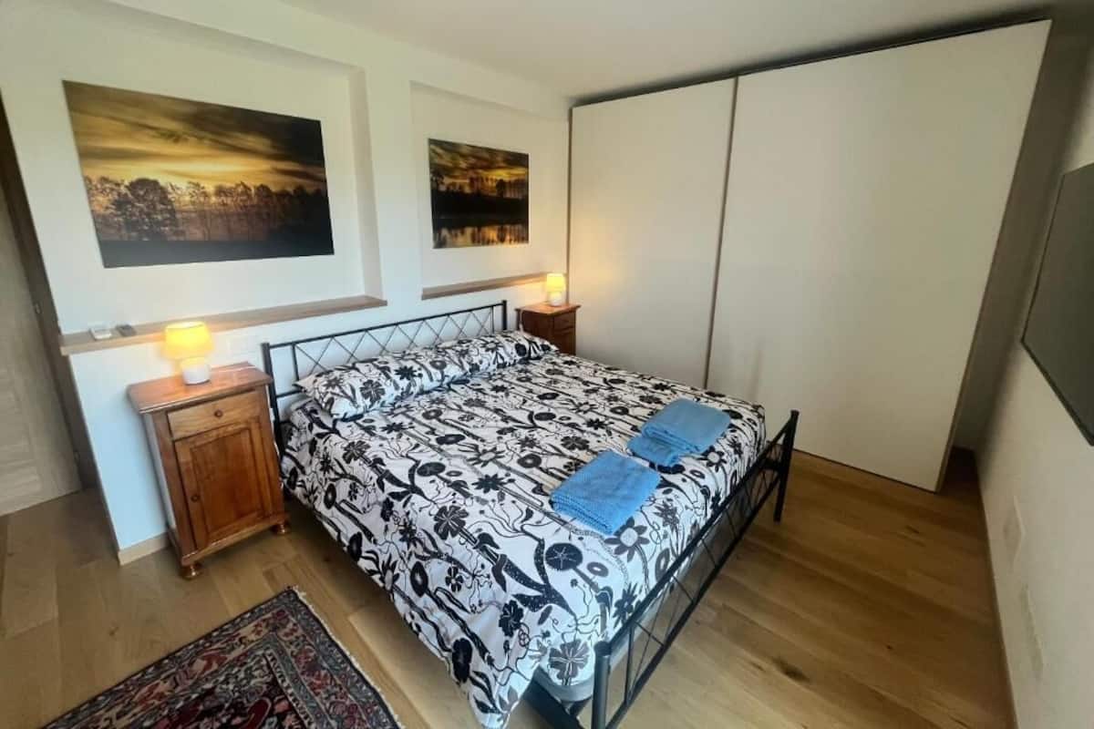 A bedroom with a bed, bedside tables, a nightstand, a rug, and two wall-mounted paintings.