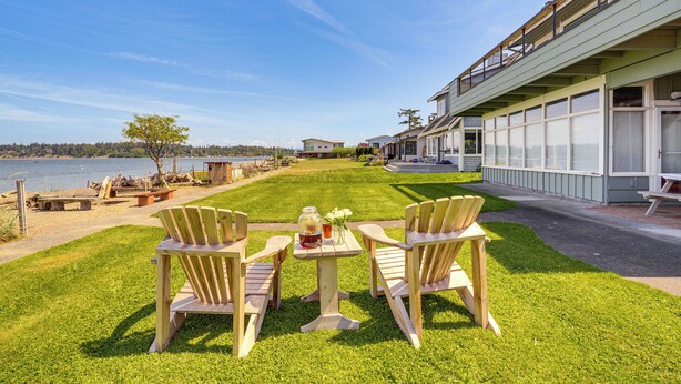 A grassy outdoor area with a table and chairs, a body of water, and a building with large windows. - Beachfront Oasis! Lummi Island Family Getaway (Lummi Island)