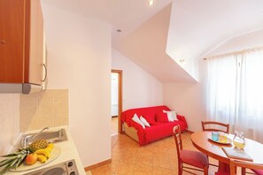 A kitchenette with a sink and stove, a dining area with a table and chairs, a red sofa, and a window with curtains. - Apartment Mondo A2 (Dubrovnik)