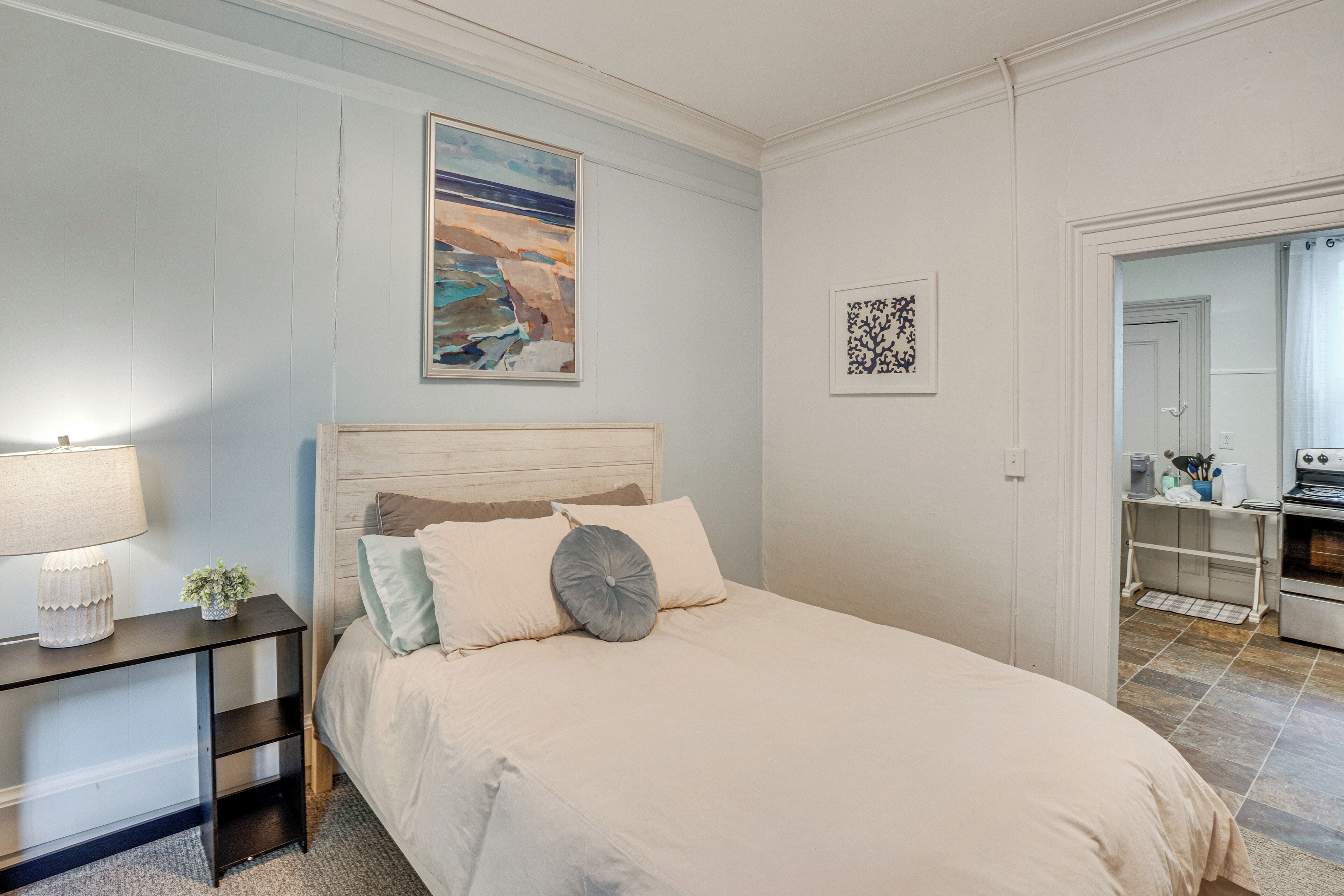 A bedroom with a bed, bedside table, lamp, and artwork on the wall.