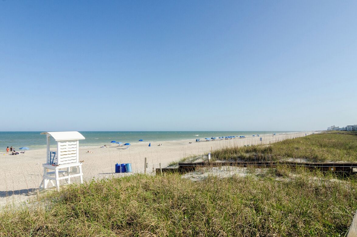 A beach with a lifeguard tower, beach umbrellas, and a grassy area in the foreground.
