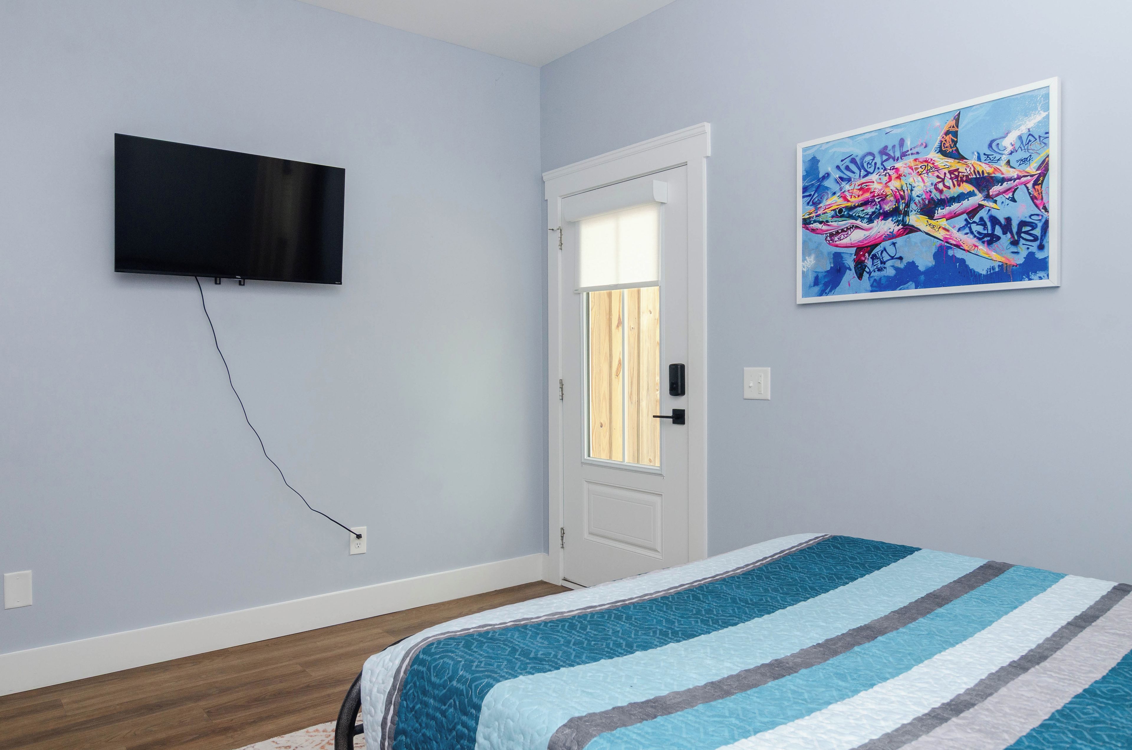 A bedroom with a bed, a television mounted on the wall, a door with a window, and a colorful painting on the wall.