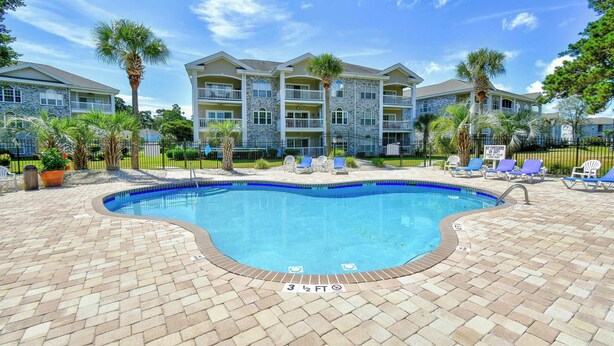 A swimming pool with a brick deck, lounge chairs, and a multi-story building in the background. - Coastal Retreat at Myrtlewood (Myrtle Beach)