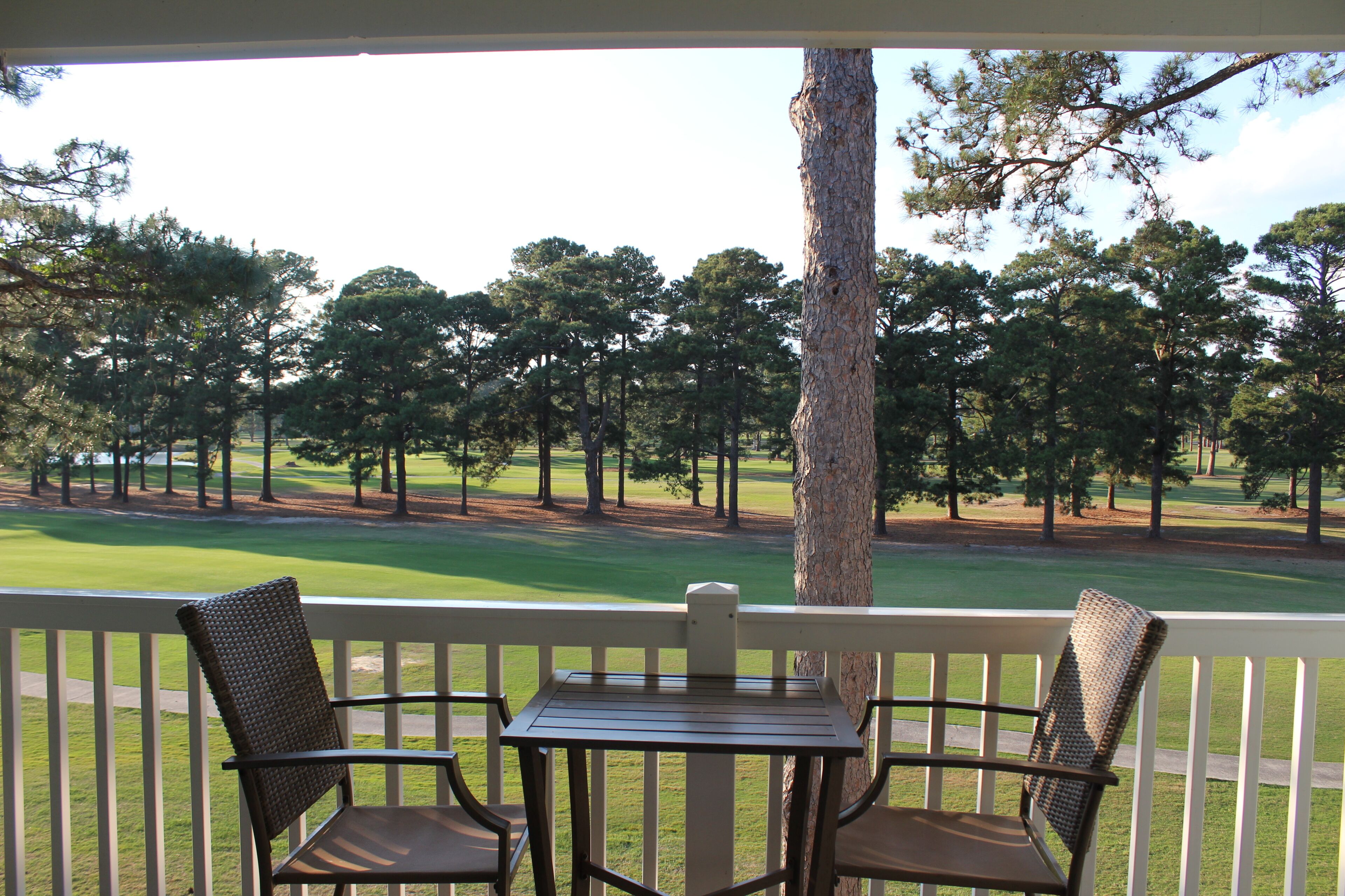 A porch with two chairs and a table, overlooking a golf course with trees and a clear sky.