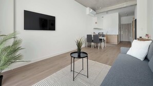 Una sala de estar moderna con un televisor de pantalla plana, un sofá gris, una pequeña mesa redonda con una planta y un área de comedor al fondo.