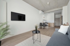Ein modernes Wohnzimmer mit Fernseher, einem grauen Sofa, einem kleinen runden Tisch mit einer Pflanze und einem Essbereich im Hintergrund.