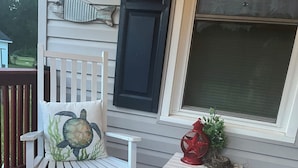 En hvid gyngestol med pude formet som skildpadde, et lille bord med en rød vase og en grå måtte på en træterrasse.