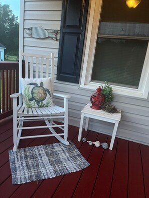 Un fauteuil à bascule blanc avec un coussin en forme de tortue, une petite table surmontée d’un vase rouge et un tapis gris sur une terrasse en bois.