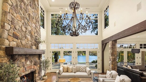 A spacious living room with a stone fireplace, a large chandelier, and a view of a lake through large windows. - Hunts Point | Waterfront Estate & Guest Villa (Hunts Point)