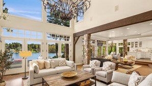 A spacious living area with a large window overlooking a lake, a fireplace, and a kitchen with white cabinets. - Hunts Point | Waterfront Estate & Guest Villa (Hunts Point)