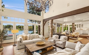 A spacious living area with a large window overlooking a lake, a fireplace, and a kitchen with white cabinets. - Hunts Point | Waterfront Estate & Guest Villa (Hunts Point)