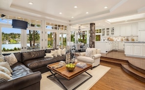 A spacious living room with a leather sofa, a coffee table with books and apples, and a view of a lake through large windows. - Hunts Point | Waterfront Estate & Guest Villa (Hunts Point)
