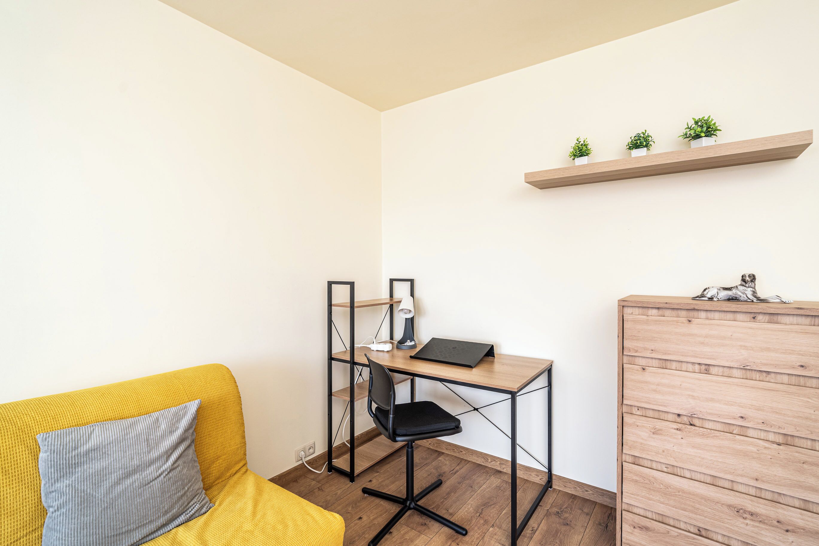Une pièce de taille modeste, avec un canapé jaune, un bureau avec une chaise, un meuble en bois et une étagère ornée de plantes.