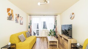 Una sala de estar con un sofá amarillo, una mesa de centro, un televisor sobre un mueble de madera y una ventana con cortinas translúcidas.