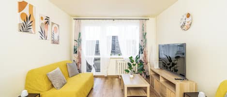 Ein Wohnzimmer mit gelbem Sofa, Couchtisch, Fernseher auf Holzfuß und Fenster mit durchsichtigen Vorhängen.