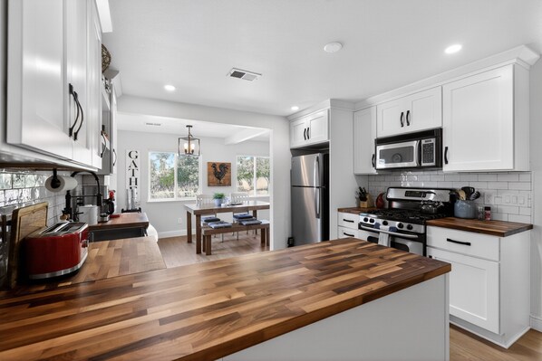 A modern kitchen with white cabinets, a wooden countertop, stainless steel appliances, and a dining area with a table and chairs. - Historic Placerville 3BR Sleeps 6 AppleHill (Placerville)