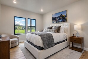 A bedroom with a large bed, a chair, a nightstand, a painting, and a view of a green landscape through large windows. - Luxury New Build W Ski Storage Fire Pit Bbq 1122 Voss 3 Bedroom Condo (Heber City)