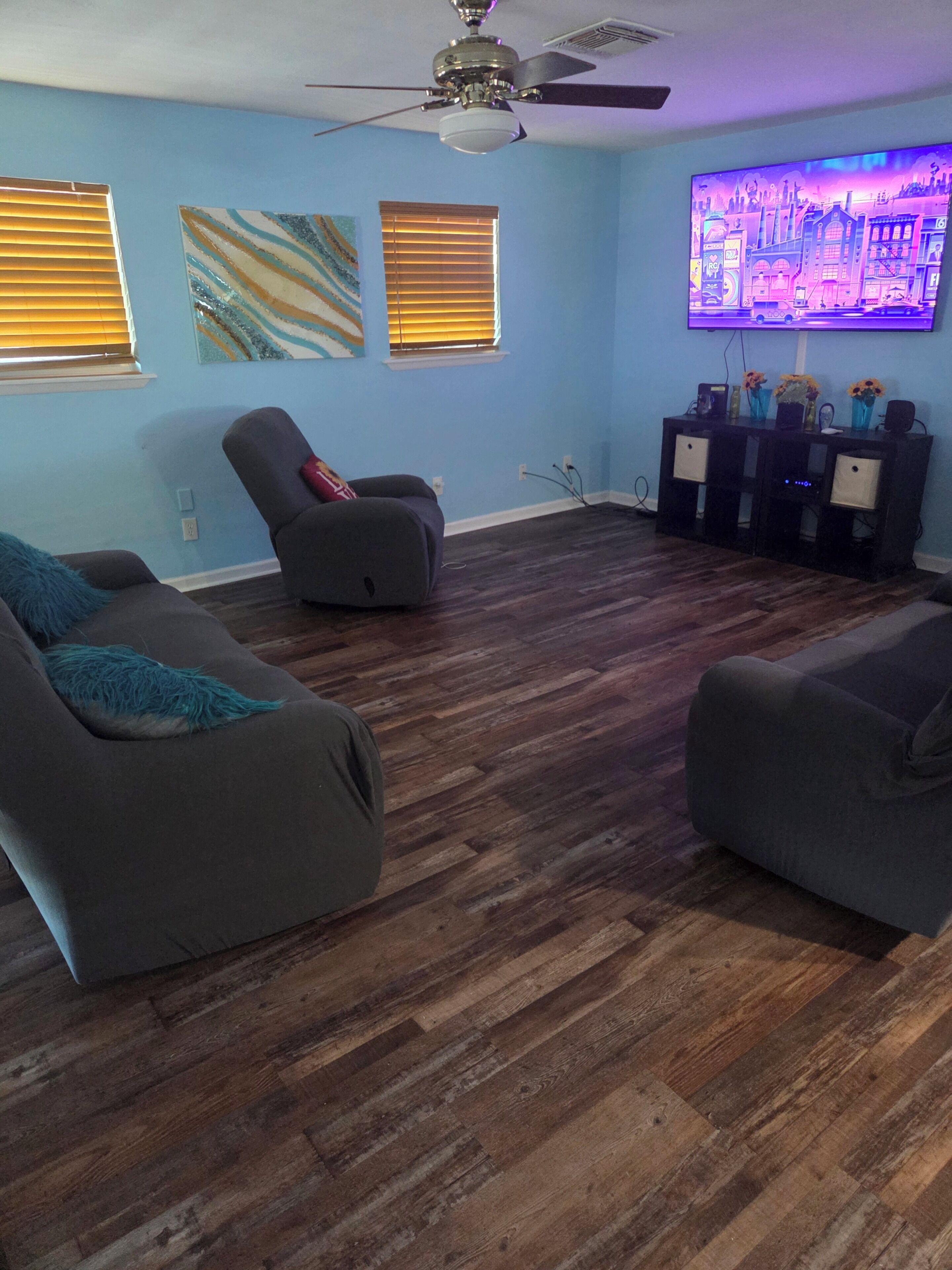 A living room with dark wood flooring, two gray sofas, a gray recliner, a ceiling fan, and a flat-screen TV mounted on the wall.