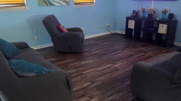 A living room with dark wood flooring, two gray sofas, a gray recliner, a ceiling fan, and a flat-screen TV mounted on the wall.