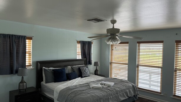 Quarto com cama, mesa de cabeceira, luminária, ventilador de teto e janelas com persianas.