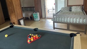 Uma mesa de bilhar com bolas, uma cama com colcha florida, uma mesinha de cabeceira com abajur e vista para o exterior.