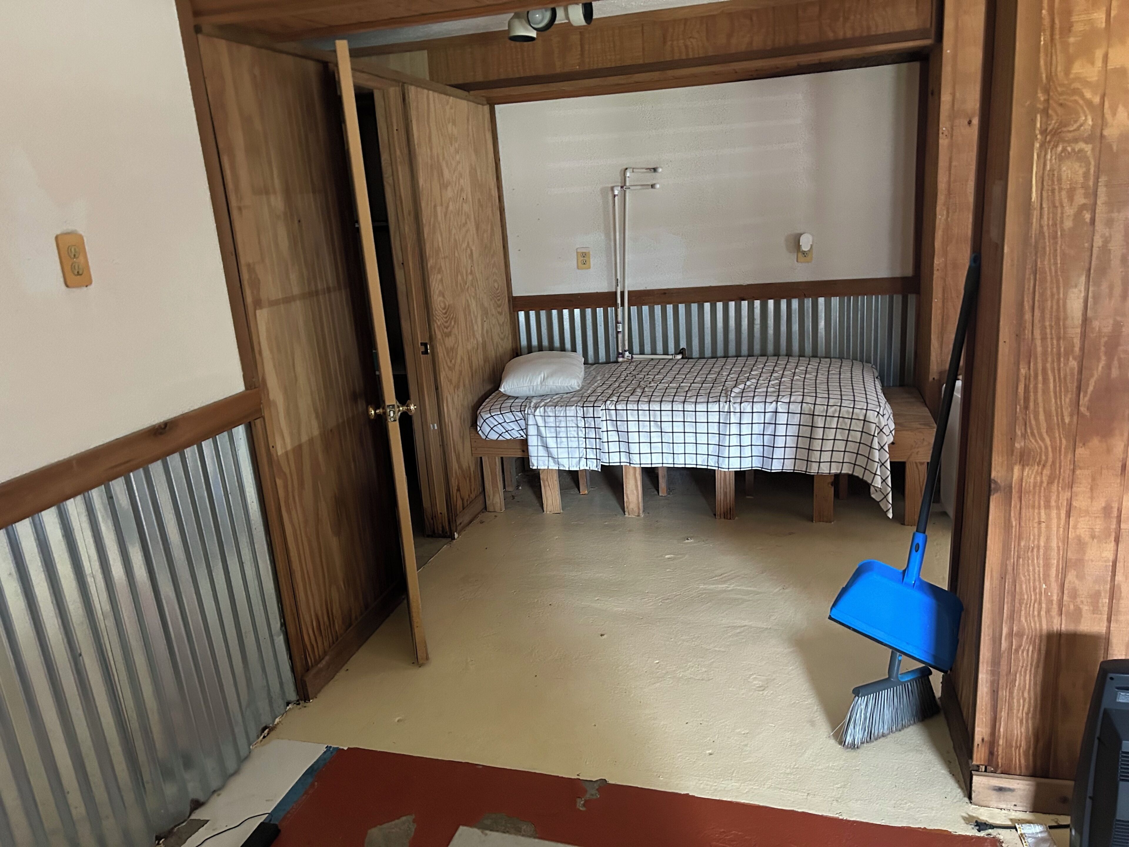 A room with a bed, a wooden wardrobe, a blue broom, and a partially visible door.