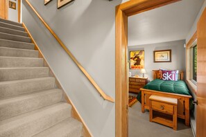 A staircase with carpeted steps and wooden handrails, a bed with a green pillow, a nightstand with a lamp, and a framed picture on the wall. - 3BR Ski-In/Out Condo, Nordic Trails, Free Shuttle Access – Big Sky, MT (Big Sky)