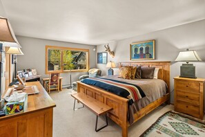 A bedroom with a wooden bed, a desk, a chair, a lamp, a bench, and a window with a view of trees. - 3BR Ski-In/Out Condo, Nordic Trails, Free Shuttle Access – Big Sky, MT (Big Sky)