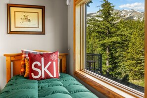 Ein Bett mit grüner Decke, ein Kopfkissen mit der Aufschrift 'SKI', ein gerahmtes Bild an der Wand und ein Fenster mit Bergblick.