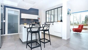 A modern kitchen with a central island, two black bar stools, and a red chair in a bright room with large windows.