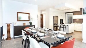A modern dining area with a long table set for six, a kitchen in the background, and a large window.