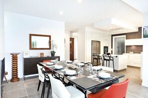 Un espace de salle à manger moderne avec une longue table dressée pour six personnes, une cuisine en arrière-plan et une grande fenêtre.