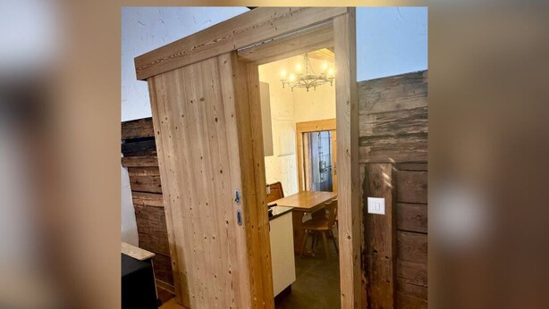 A wooden sliding door leading to a room with a dining table and chairs, a chandelier, and a small kitchenette.