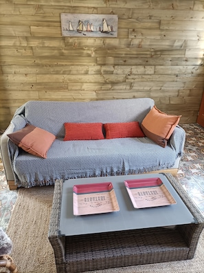 Un mur en bois orné d’un tableau représentant des voiliers, un canapé à motifs avec des coussins rouges et une table basse en osier sur laquelle reposent deux plateaux.
