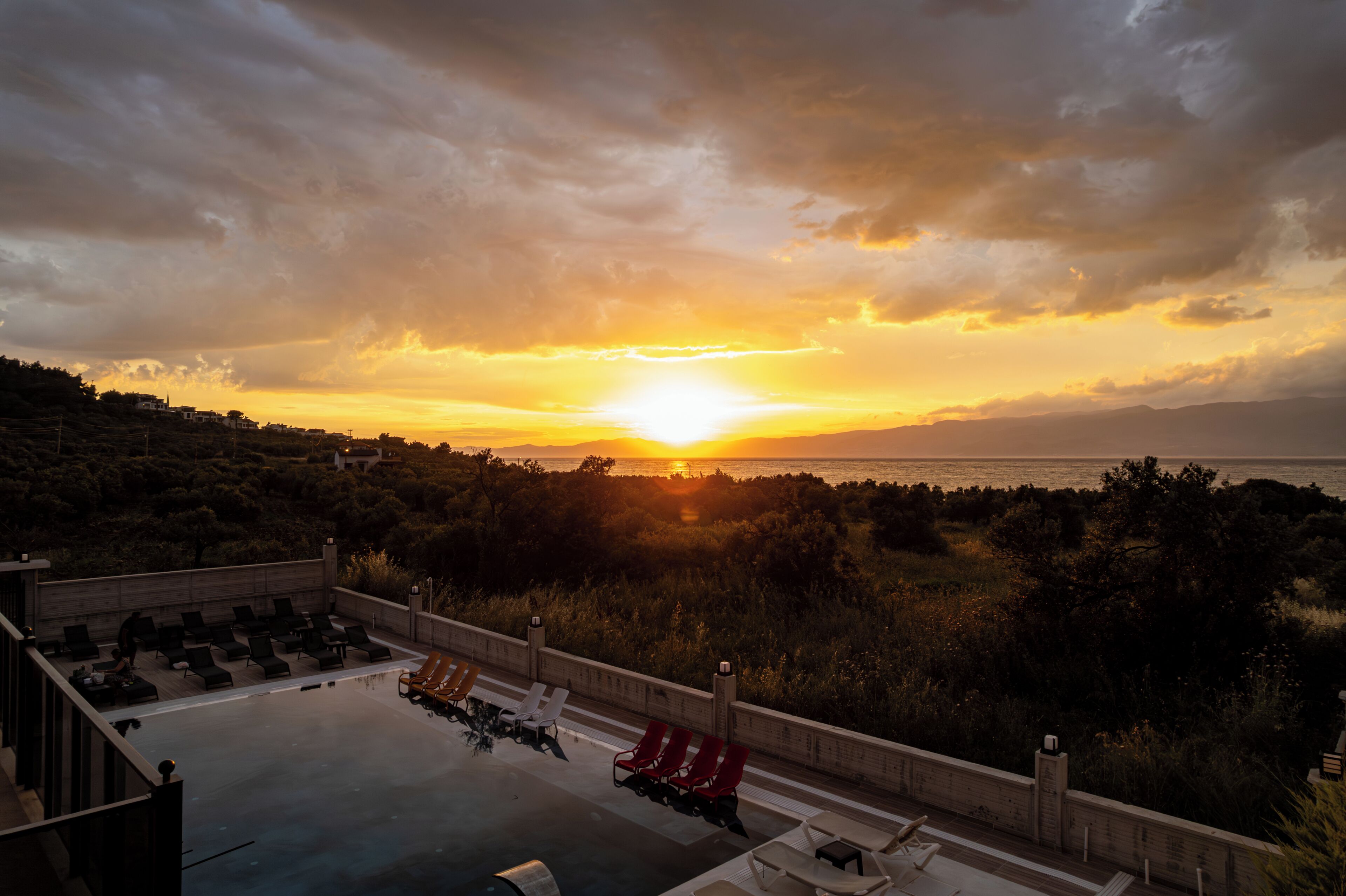 Una piscina en la azotea con sillones de descanso, vista al atardecer sobre el mar y un paisaje montañoso.