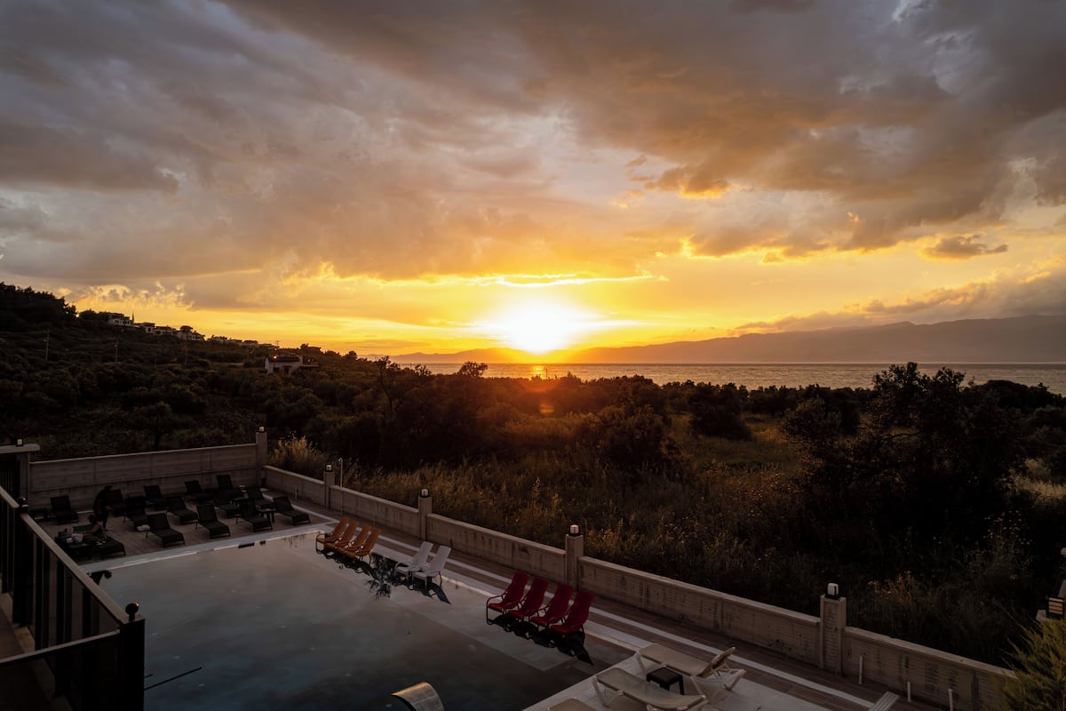 Una piscina en la azotea con sillones de descanso, vista al atardecer sobre el mar y un paisaje montañoso.
