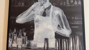 Ein gerahmtes Kunstwerk mit einer skelettartigen Figur, die ein Getränk hält, mit einer Bar und Flaschen im Hintergrund.