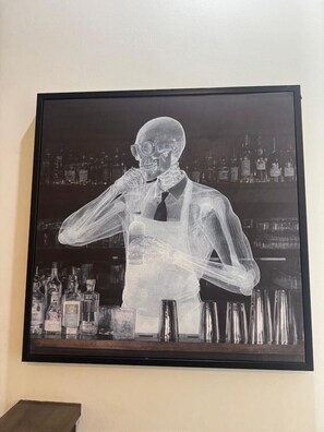 Une œuvre d’art encadrée représentant une figure squelettique tenant un verre, avec un bar et des bouteilles en arrière-plan.