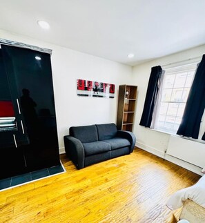 A living room with a black sofa, a wooden cabinet, a painting on the wall, and a window with curtains. - Cosy one bedroom flat near Aldgate East station (Greater London)