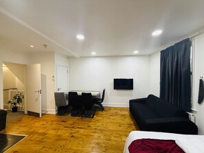 A room with a dining table, chairs, a black sofa, a TV mounted on the wall, and a window with curtains. - Wonderful one bedroom apartment in Great location (Greater London)