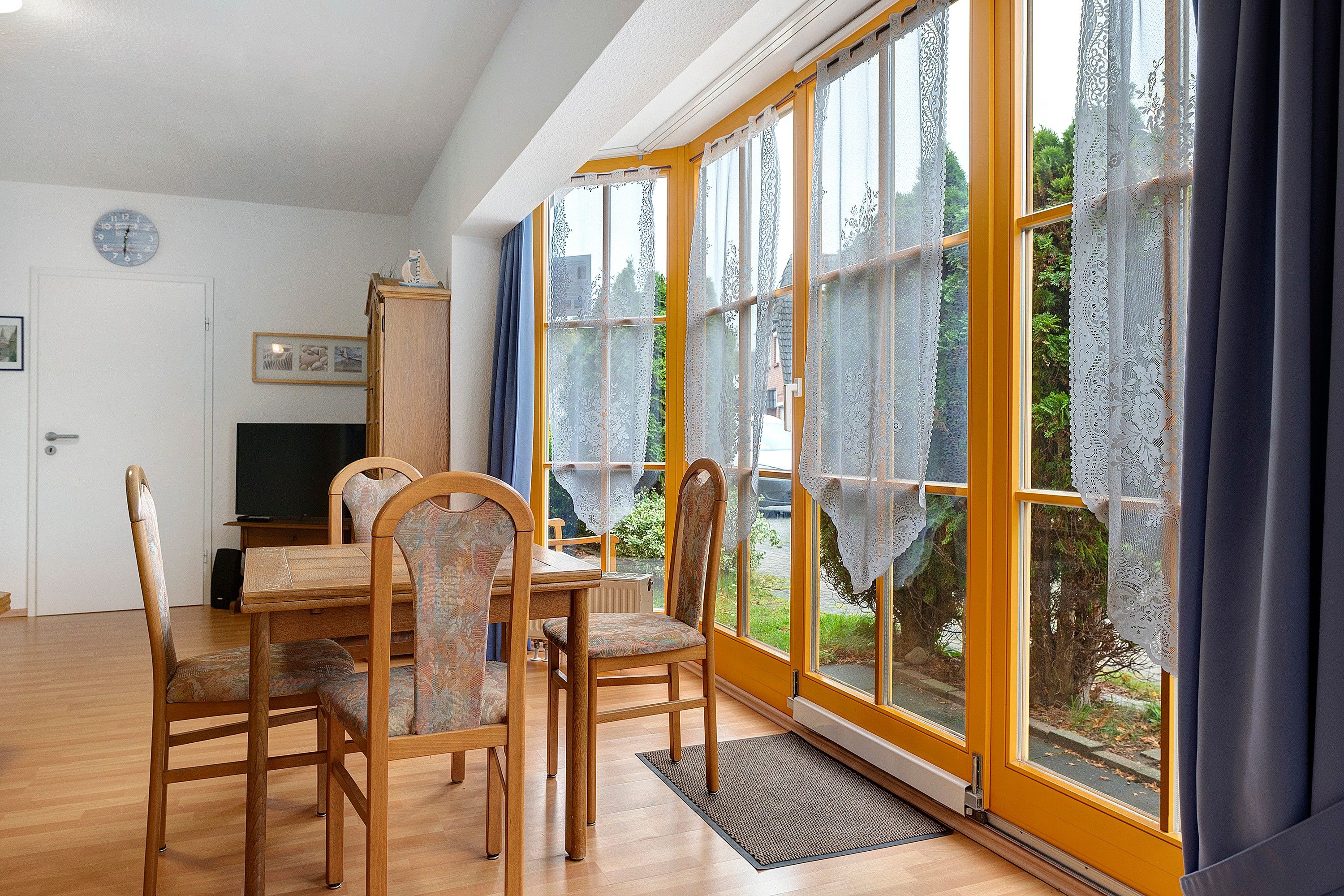 Ein Essbereich mit Tisch, Stühlen, einem Fernseher und einem Fenster mit Spitzenvorhängen.