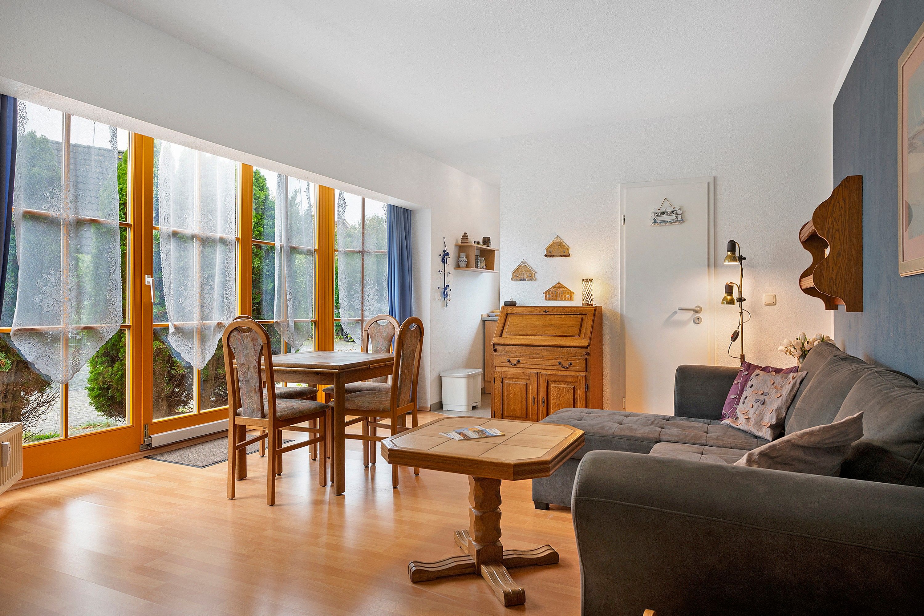 Ein Wohnzimmer mit einem Holzesstisch, Stühlen, einer Couch und einem Couchtisch. Es gibt ein großes Fenster mit Vorhängen, eine Kommode und Wanddekorationen.