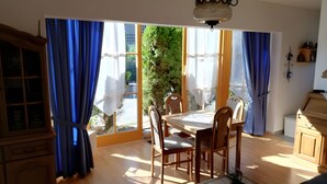 Ein Essbereich mit Tisch und Stühlen, großen Fenstern mit Vorhängen und Blick auf Grünflächen.