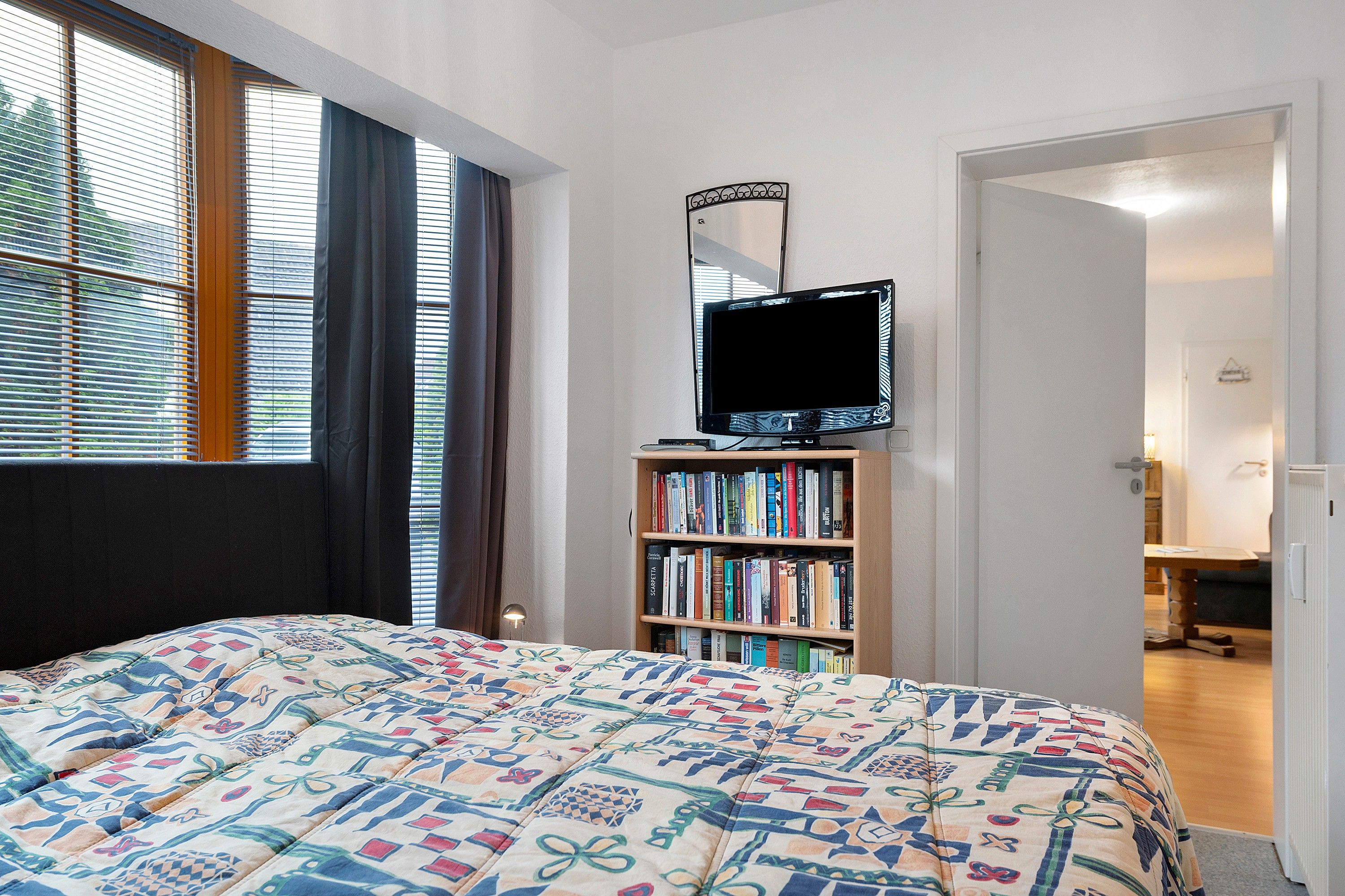 Ein Schlafzimmer mit einem Bett, einem Fernseher auf einem Schrank, einem Bücherregal und einem Fenster mit Jalousien.