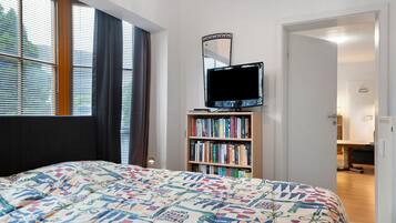 Ein Schlafzimmer mit einem Bett, einem Fernseher auf einem Schrank, einem Bücherregal und einem Fenster mit Jalousien.