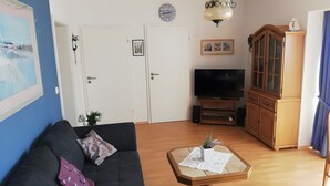 Ein Wohnzimmer mit einer schwarzen Couch, einem Couchtisch mit einer Zeitung, einem Fernseher und einer Vitrine mit Glastüren.