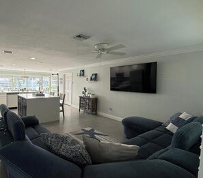 Ein modernes Wohnzimmer mit großem Flachbildfernseher, dunkelblauen Sofas, einem Essbereich und einer Küche im Hintergrund sowie einem Deckenventilator.