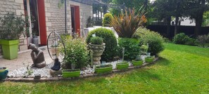 Un edificio de piedra con una puerta roja, un jardín con diversas plantas y una estatua, y un césped bien cuidado.