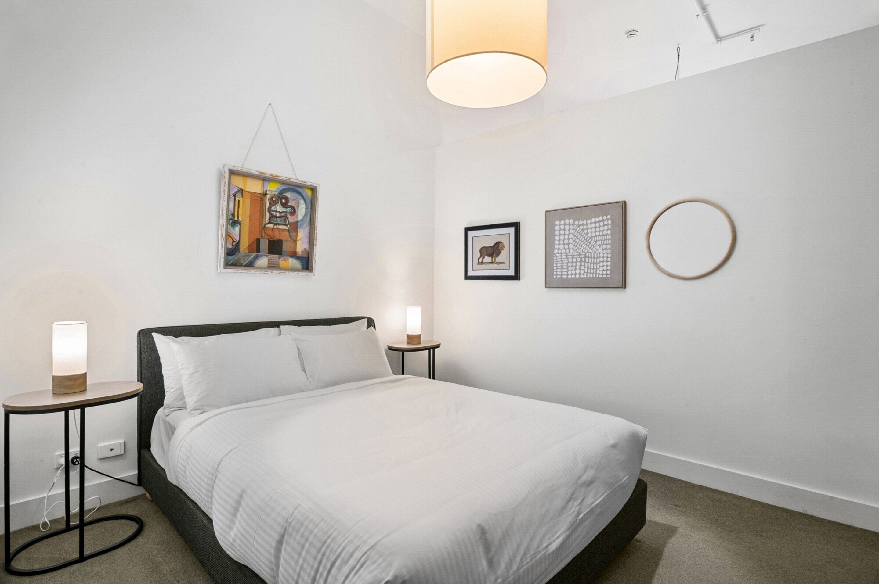 A neatly arranged hotel room with a bed, bedside tables, a hanging artwork, and a wall-mounted light fixture.