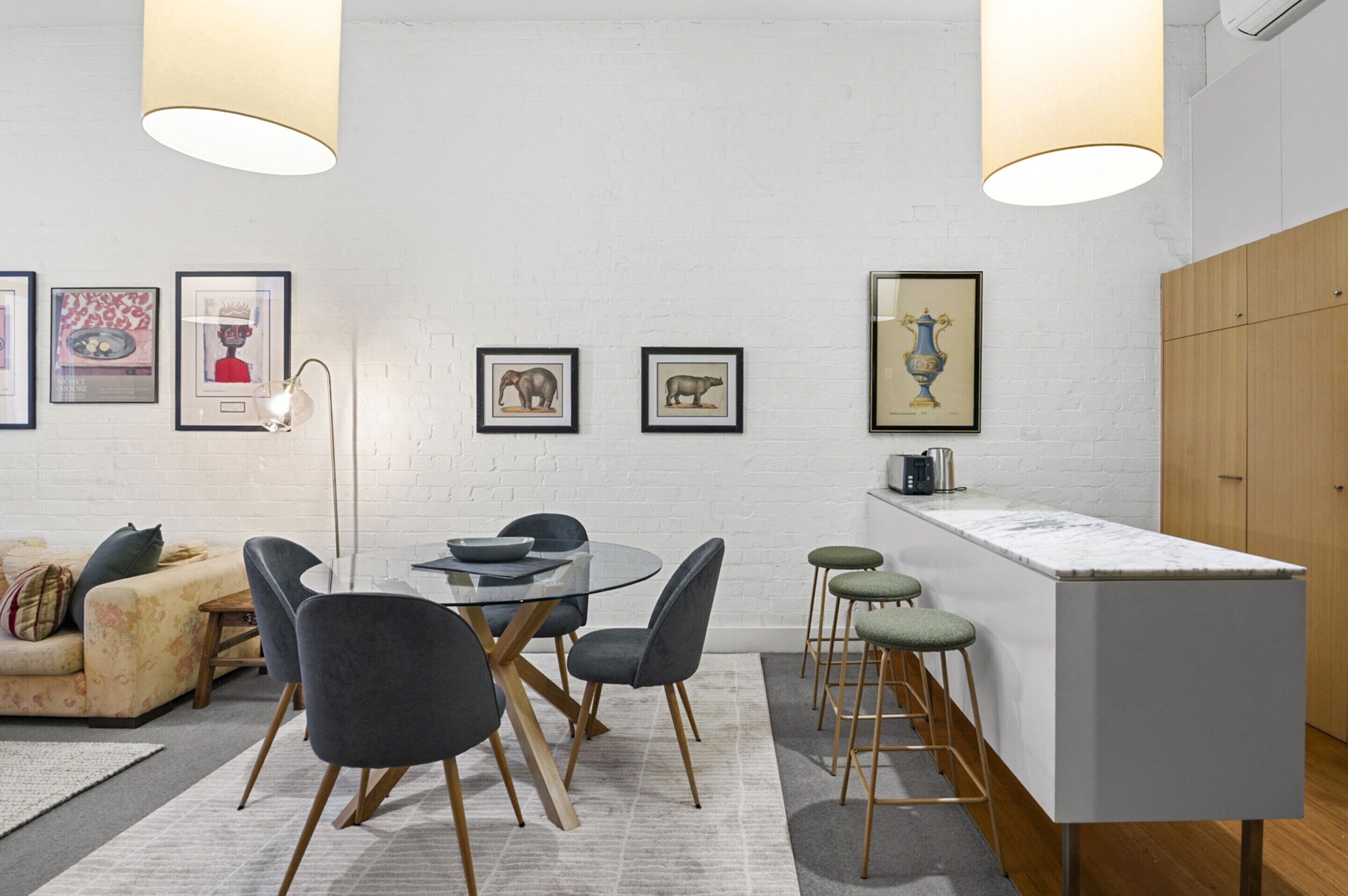 A modern kitchen with a white brick wall, a round dining table with chairs, a bar area with stools, and framed pictures on the wall.